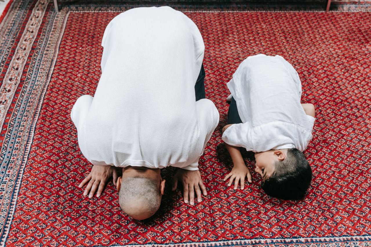 A father and son kneeling in prayer on a beautifully patterned red carpet, symbolizing religious devotion.