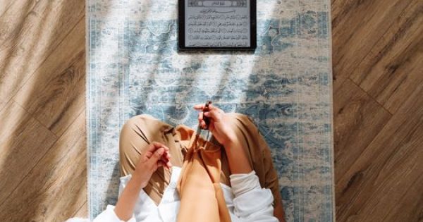 High-angle view of a person praying with prayer beads and Quran on a rug.