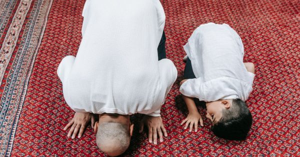 A father and son kneeling in prayer on a beautifully patterned red carpet, symbolizing religious devotion.